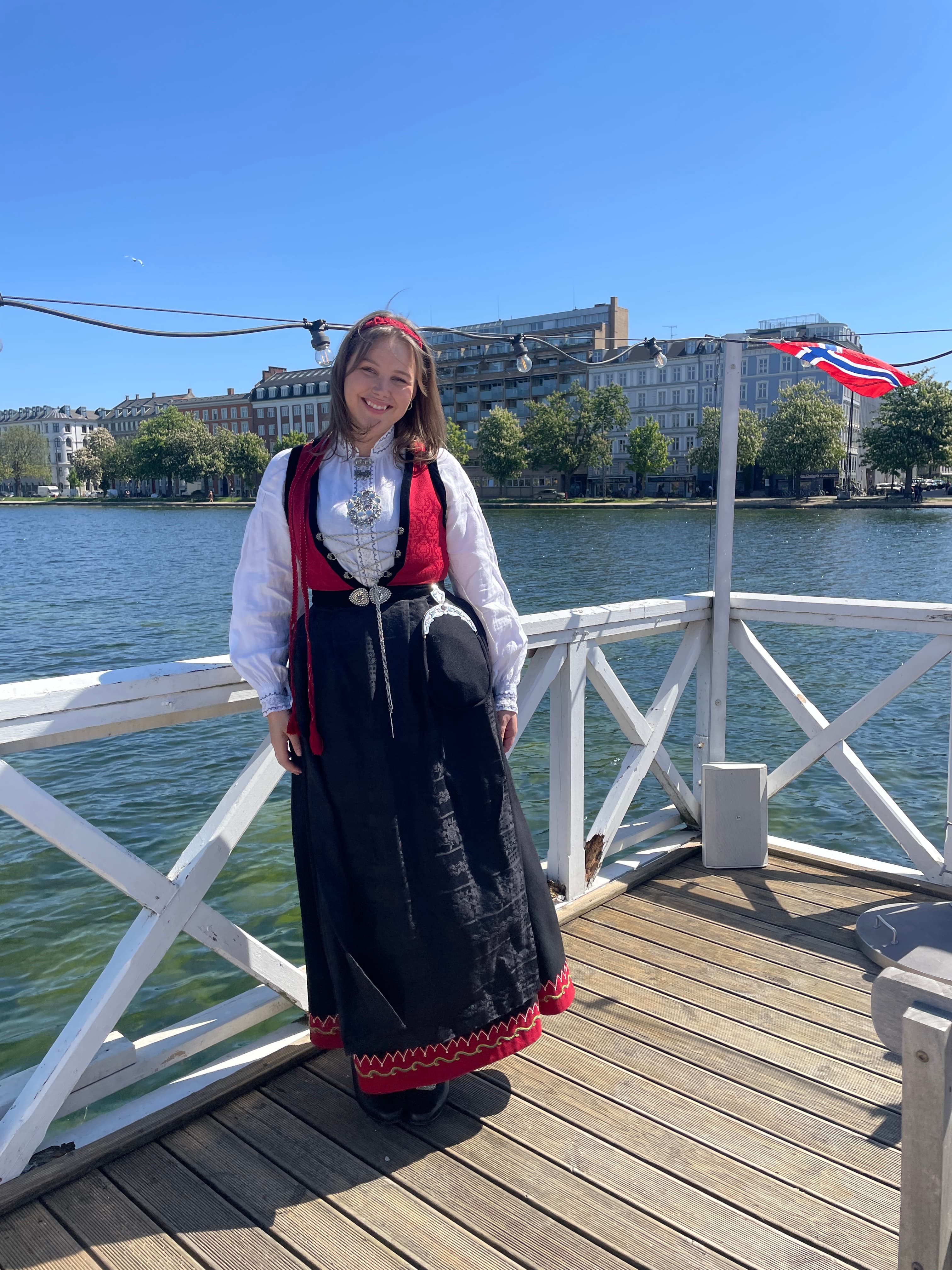 Standing by the waterfront in traditional Norwegian bunad, with a Norwegian flag