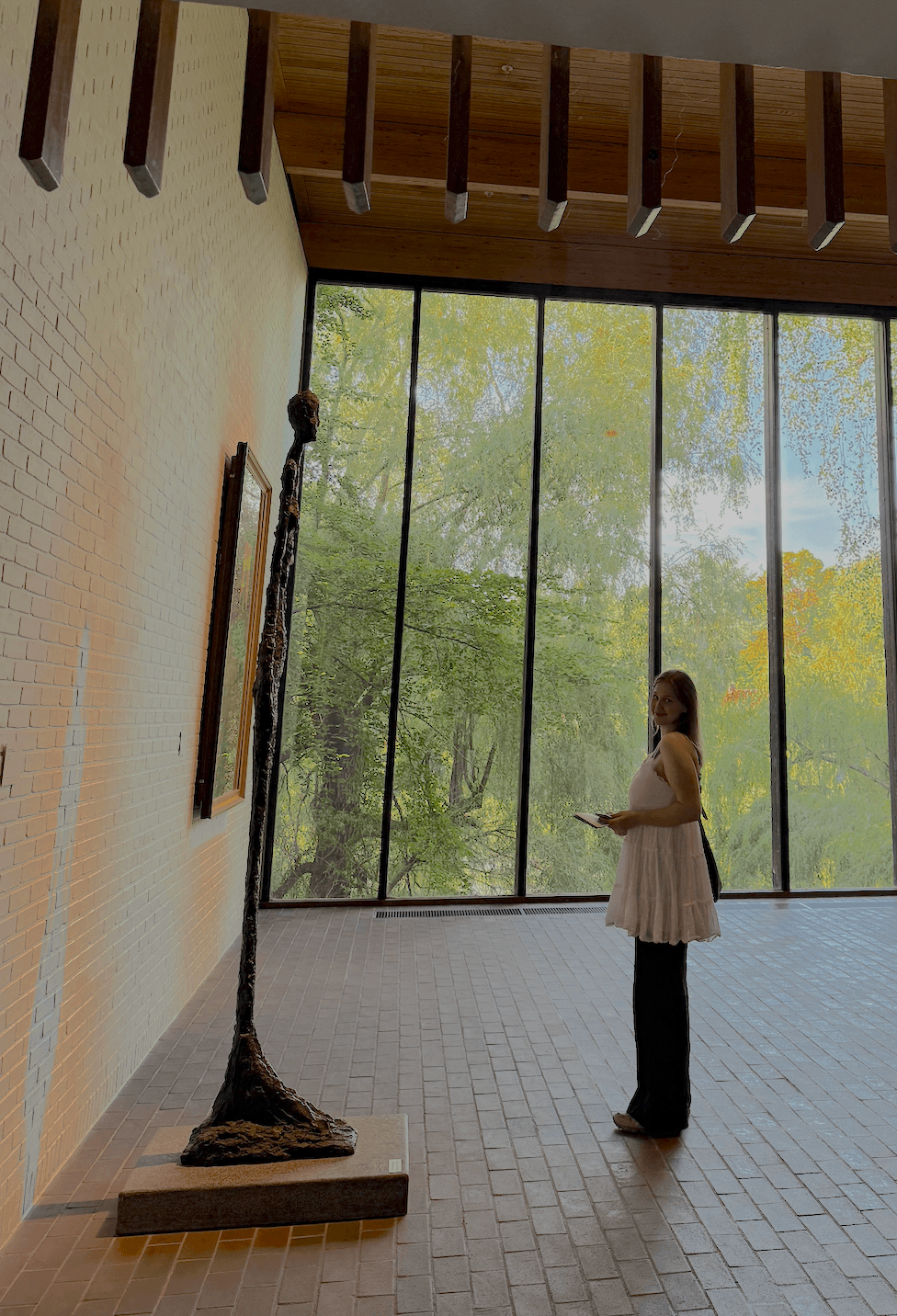 Emma standing in a gallery, looking at a sculpture beside a wall of windows overlooking trees